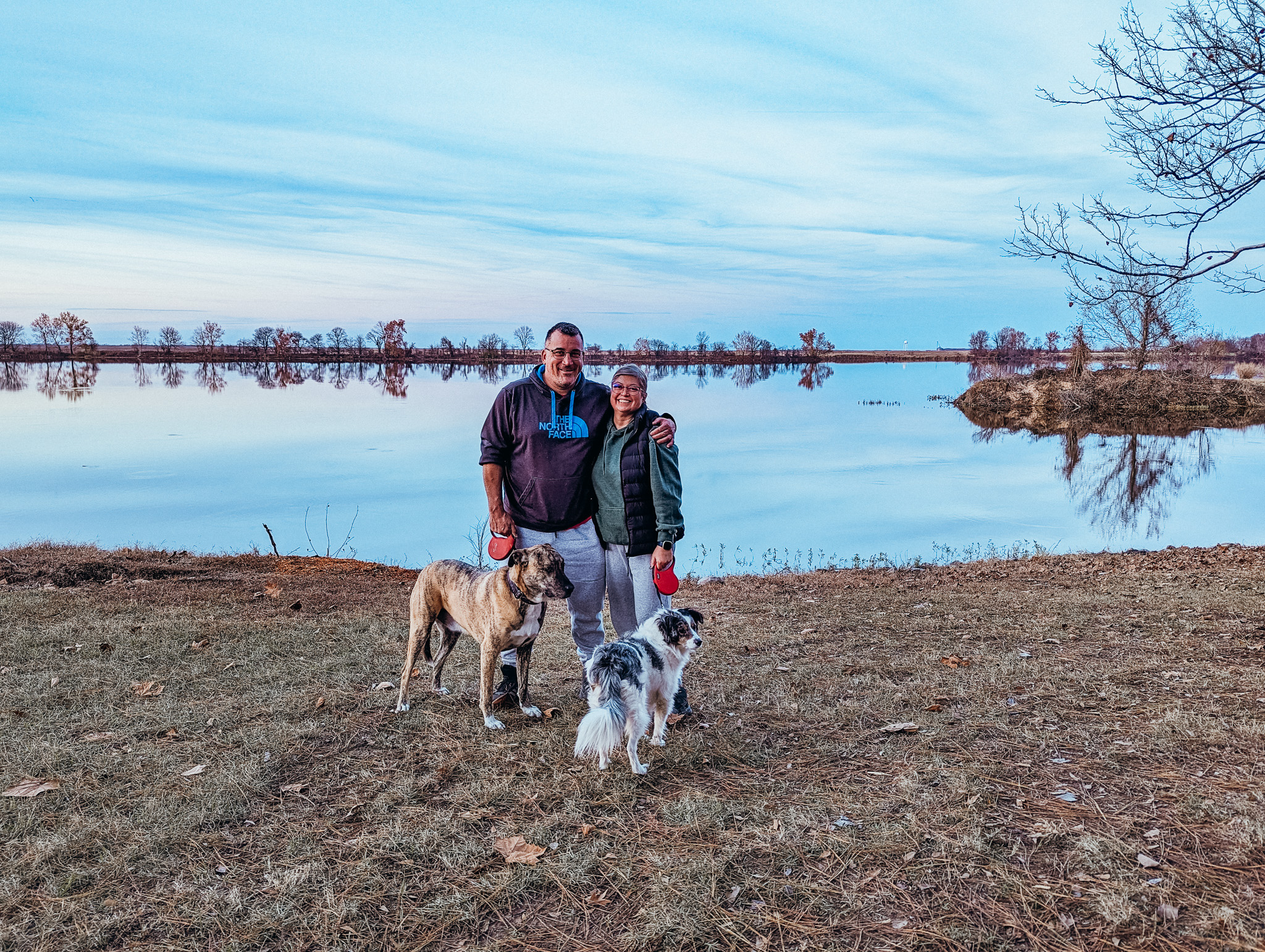 Man and woman smiling at camera with their two dogs. Behind them is a lake at a state park. Picture taken at sunset and the blue sky is reflecting in the water.